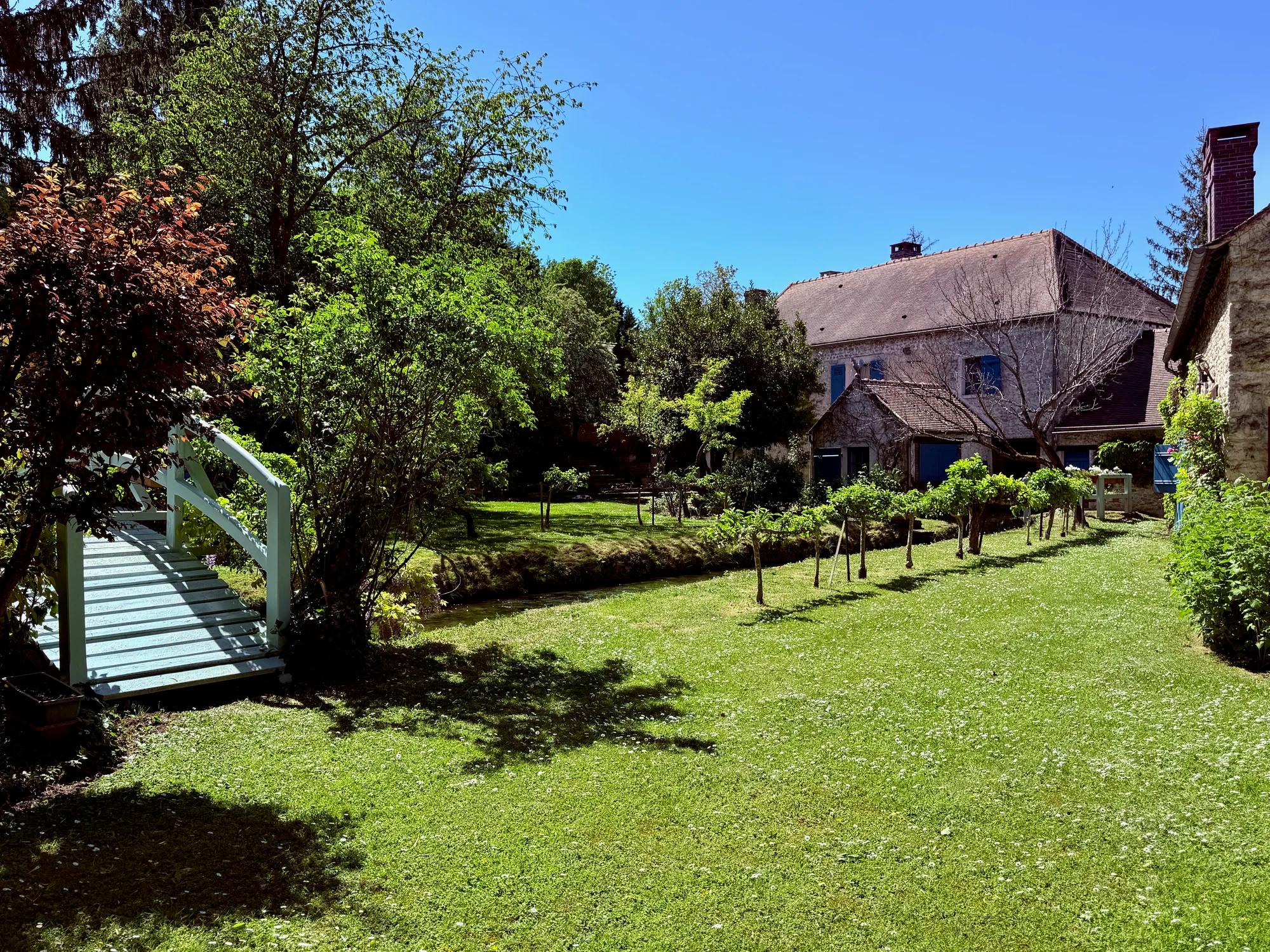 The broad garden lawn with the white footbridge and espaliered fruit trees at Moulin à Rêves