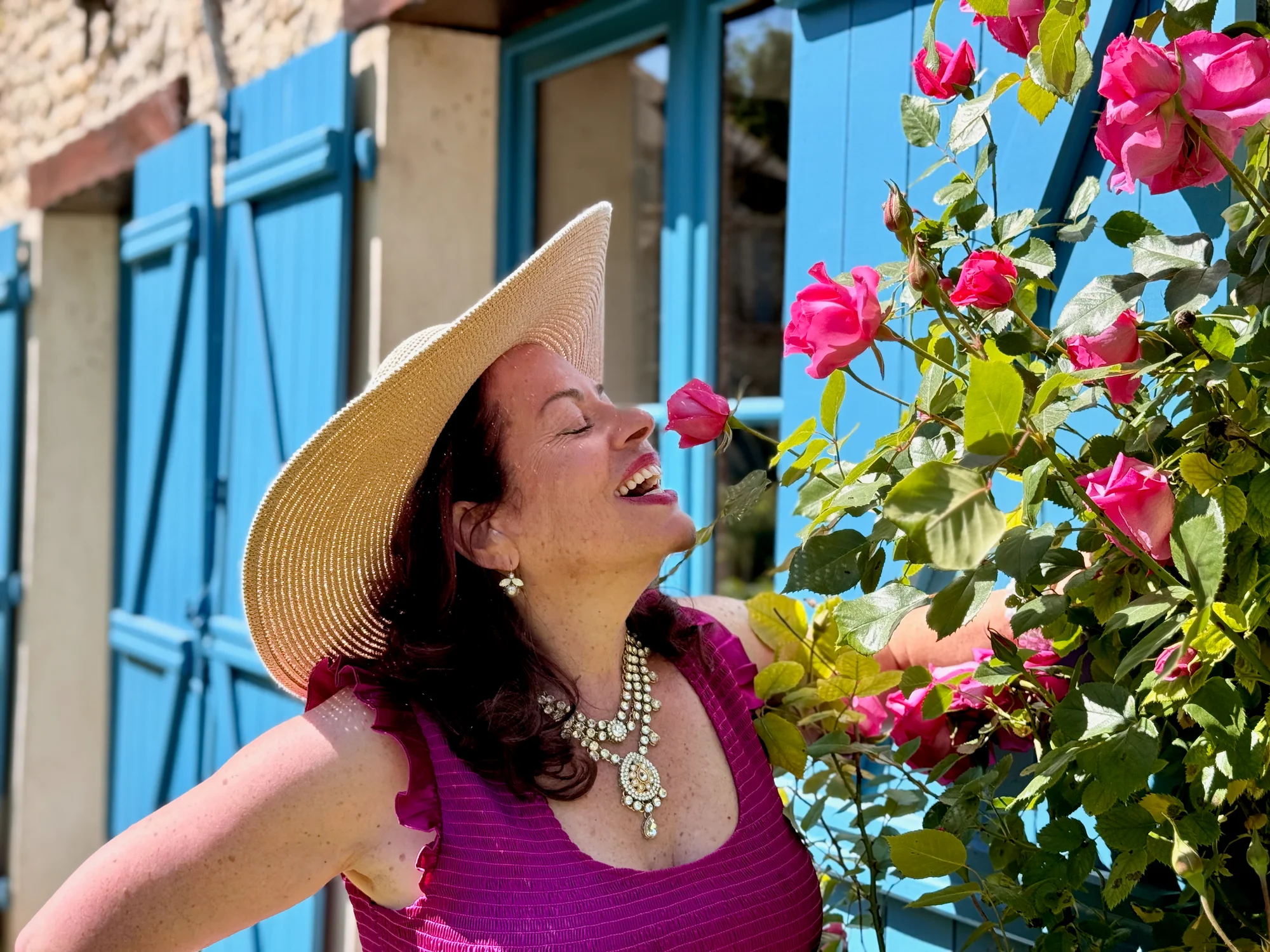 A guest in a sun hat leaning in to smell the pink roses against the blue shutters at Moulin à Rêves
