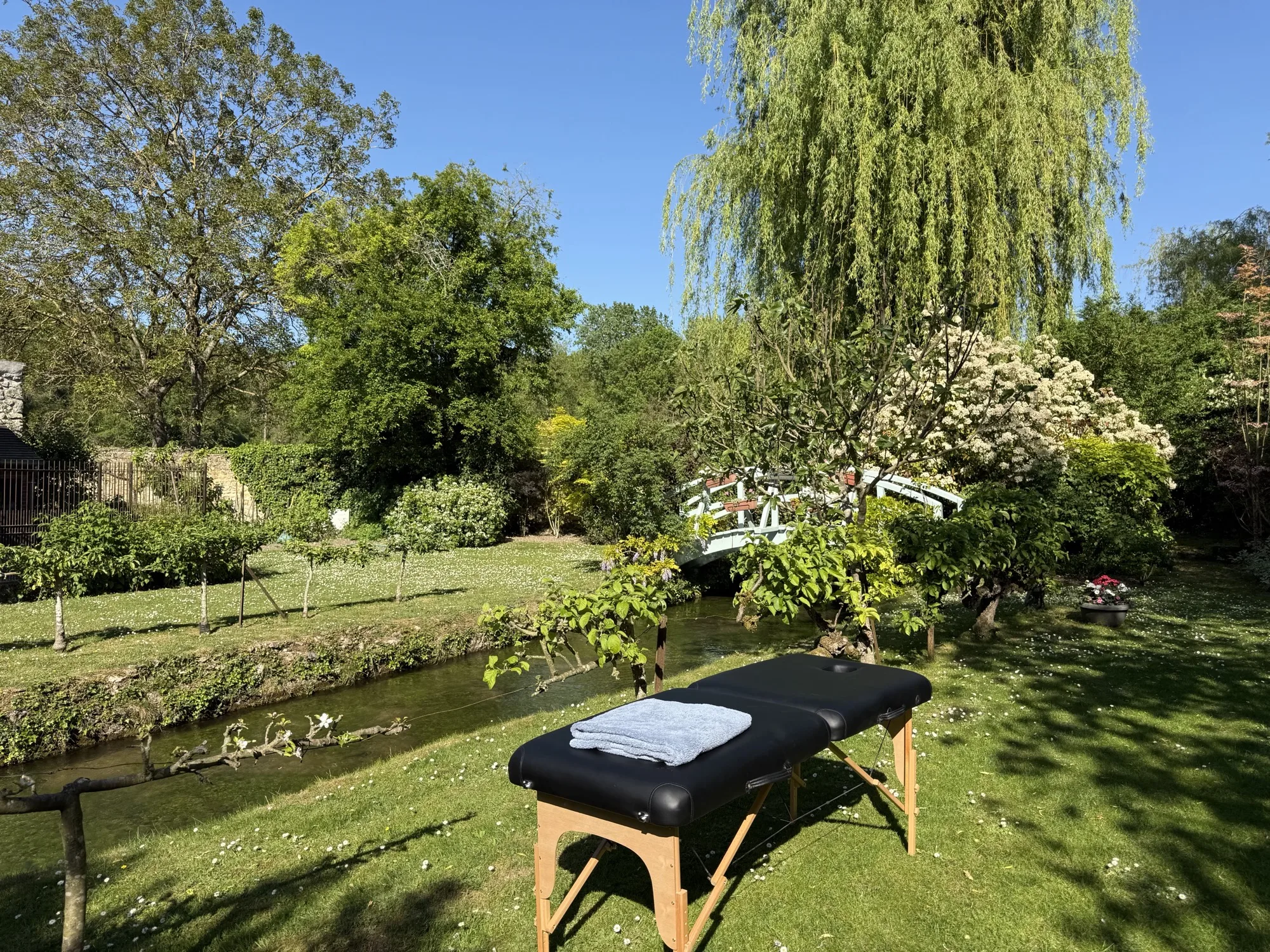 Massage table on the lawn beneath the willow, with the painted bridge beyond, at Moulin à Rêves