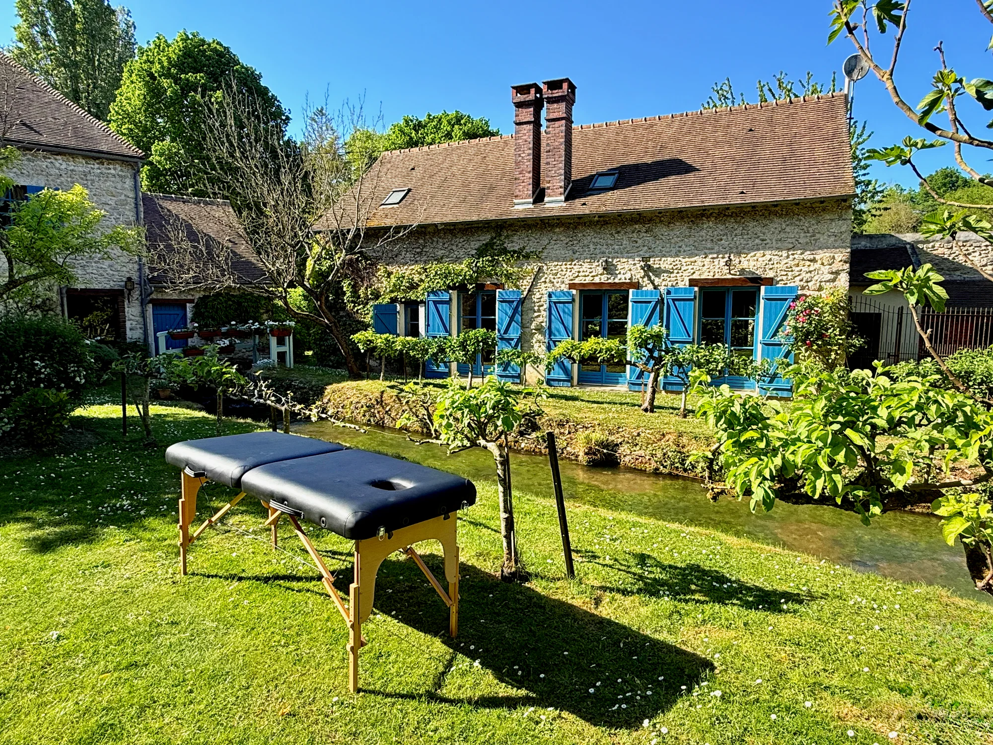 Massage table on the sunlit lawn beside the millstream and blue-shuttered mill house